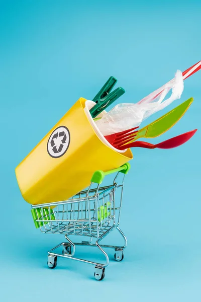 Papelera de reciclaje amarillo con plástico en un pequeño carrito de compras sobre fondo azul - foto de stock