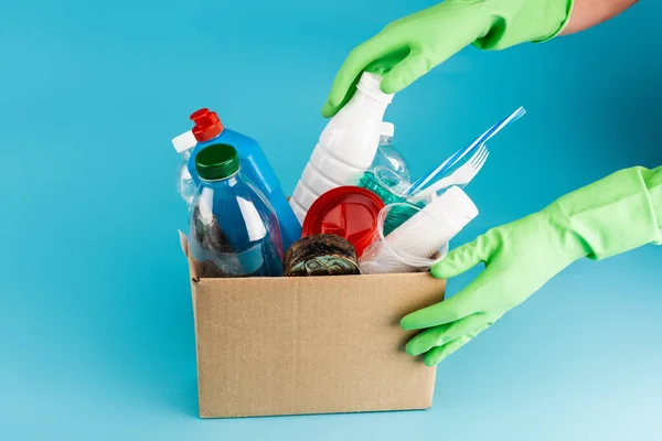 Vista recortada de limpiador en guantes de goma recogiendo basura en caja de cartón sobre fondo azul - foto de stock