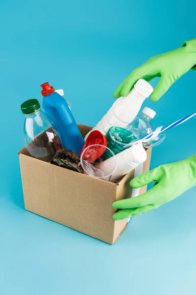 Vue recadrée du nettoyant dans des gants en caoutchouc ramassant les déchets dans une boîte en carton sur fond bleu — Photo de stock