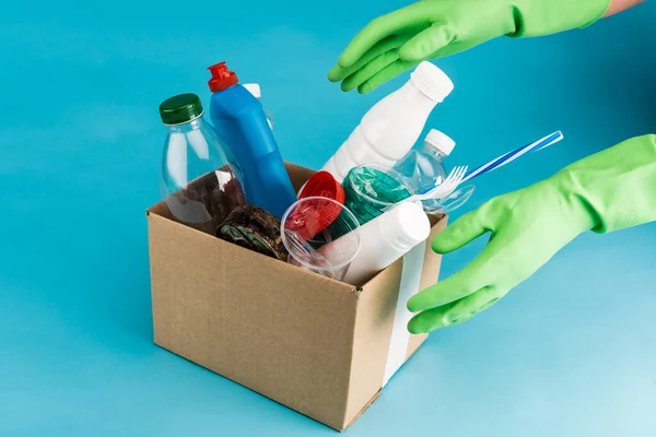 Vista recortada de limpiador en guantes de goma recogiendo basura en caja de cartón sobre fondo azul - foto de stock