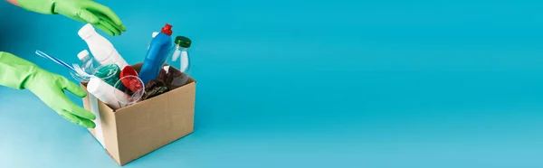 Cropped view of cleaner in rubber gloves collecting rubbish in cardboard box on blue background, panoramic shot — Stock Photo