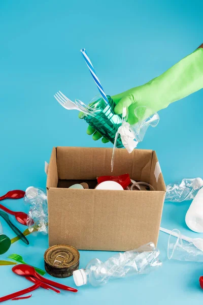 Cropped view of cleaner in rubber glove collecting rubbish in cardboard box on blue background — Stock Photo