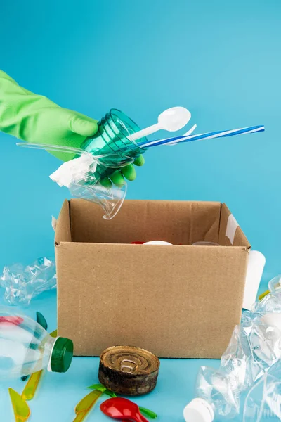 Abgeschnittene Ansicht eines Reinigers im Gummihandschuh, der Müll in einem Karton auf blauem Hintergrund sammelt — Stockfoto