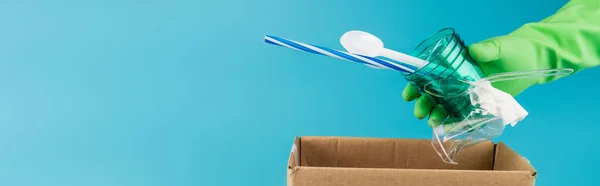 Cropped view of cleaner in rubber glove collecting plastic rubbish in cardboard box isolated on blue, panoramic shot — Stock Photo