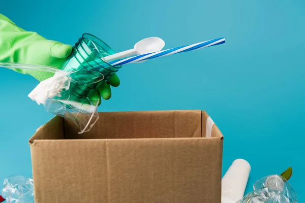 Abgeschnittene Ansicht eines Reinigers im Gummihandschuh, der Plastikmüll in einem Karton auf blauem Hintergrund sammelt — Stockfoto