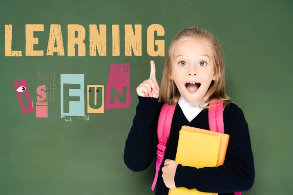 Excited schoolgirl holding book and showing idea gesture near green chalkboard with learning is fun illustration — Stock Photo
