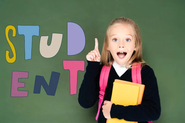 Excited schoolgirl holding book and showing idea gesture while standing near green chalkboard with student illustration — Stock Photo