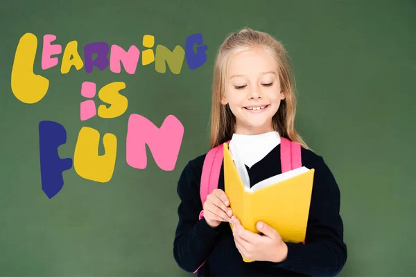 Schoolgirl reading book near green chalkboard with learning is fun illustration — Stock Photo