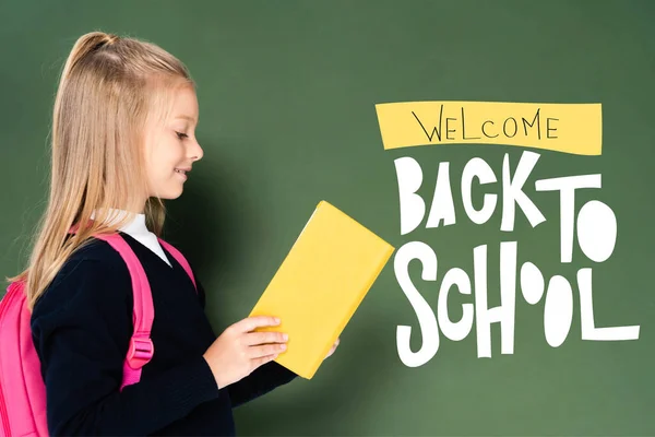 Side view of schoolgirl reading book near green chalkboard with welcome back to school illustration — Stock Photo