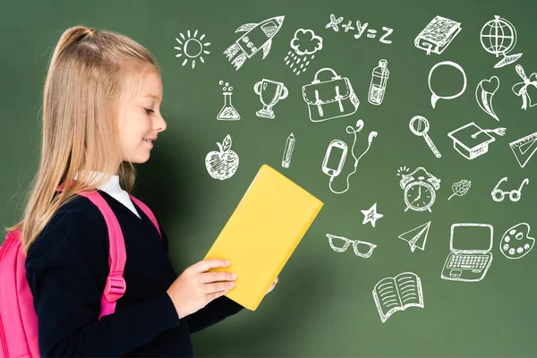 Side view of schoolgirl reading book near green chalkboard with educational illustration — Stock Photo