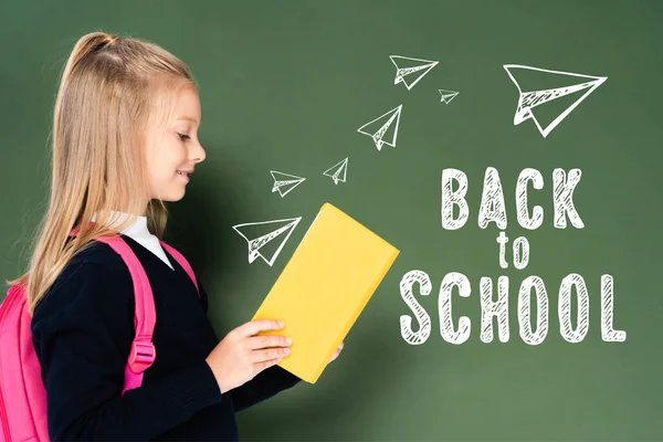 Side view of schoolgirl reading book near green chalkboard with back to school illustration — Stock Photo