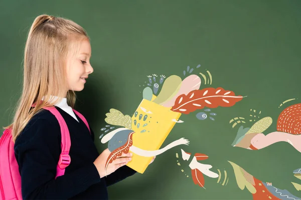 Side view of schoolgirl reading book with fairy illustration near green chalkboard — Stock Photo