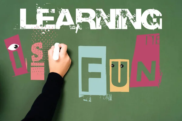 Cropped view of schoolgirl writing with chalk learning is fun lettering on green chalkboard — Stock Photo