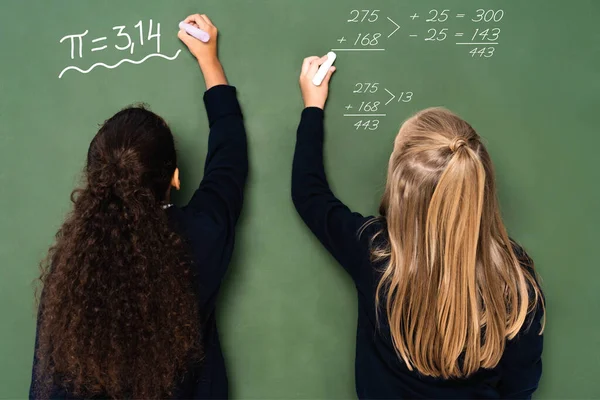 Vue arrière des écolières multiculturelles écrivant des équations mathématiques sur tableau noir — Photo de stock
