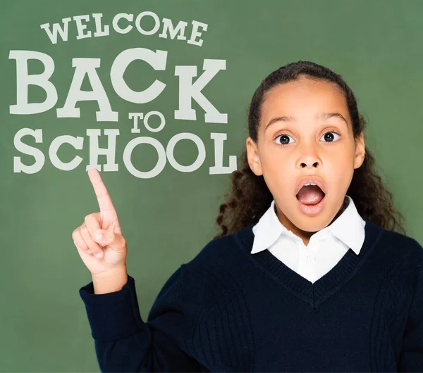 Shocked african american schoolgirl pointing at back to school lettering on green chalkboard — Stock Photo