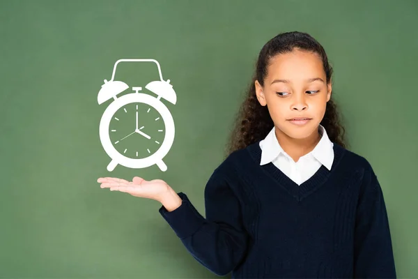 Скептически настроенная школьница указывает рукой на будильник на зеленой доске — стоковое фото