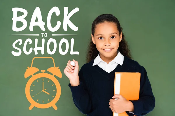 Afro-américaine écolière tenant morceau de craie et de livre tout en se tenant près de tableau noir avec retour à l'école lettrage — Photo de stock