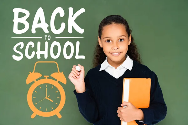 Colegiala afroamericana sosteniendo pedazo de tiza y libro mientras está de pie cerca de pizarra con letras de vuelta a la escuela - foto de stock