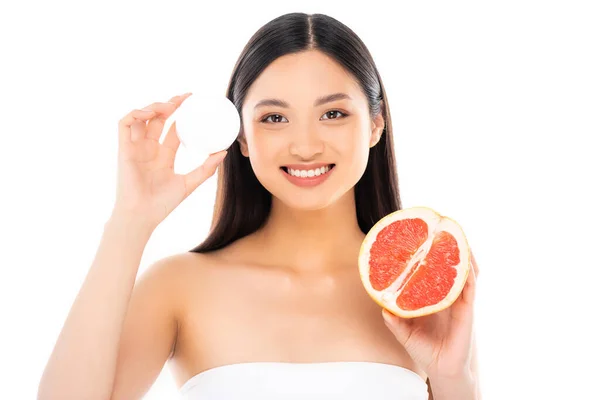 Morena mujer asiática mirando a cámara mientras sostiene la mitad de jugosa pomelo y crema cosmética aislado en blanco - foto de stock