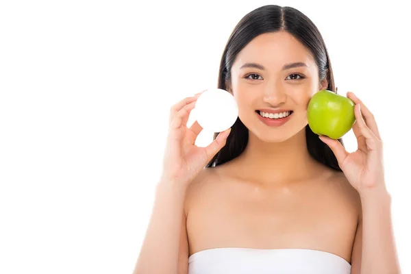 Брюнетка азіатська жінка тримає свіже зелене яблуко і косметичний крем, дивлячись на камеру ізольовано на білому — стокове фото