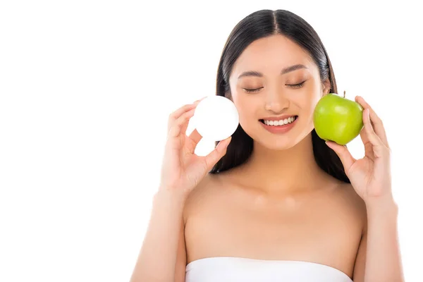 Jeune femme asiatique avec les yeux fermés tenant crème cosmétique et pomme verte entière isolé sur blanc — Photo de stock