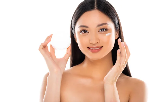 Mujer asiática desnuda mirando a la cámara mientras que la aplicación de crema cosmética en la cara aislada en blanco - foto de stock