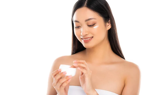 Brunette asian woman touching cosmetic cream while applying it on face isolated on white — Stock Photo