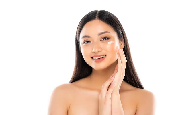 Mujer asiática desnuda aplicando crema cosmética en la cara mientras mira la cámara aislada en blanco - foto de stock