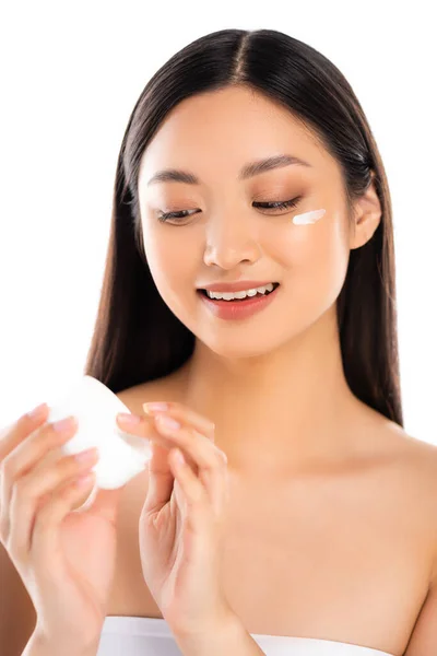 Jeune femme asiatique toucher la crème cosmétique tout en l'appliquant sur le visage isolé sur blanc — Photo de stock