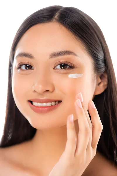 Brünette asiatische Frau, die in die Kamera schaut, während sie kosmetische Creme auf das Gesicht aufträgt, isoliert auf weiß — Stockfoto