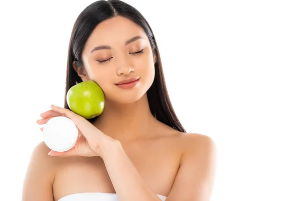 Молода азіатка з закритими очима тримає зелене яблуко і косметичний крем біля обличчя ізольовані на білому — стокове фото