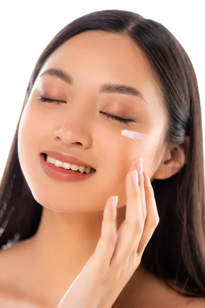 Morena mujer asiática con los ojos cerrados aplicando crema cosmética en la cara aislada en blanco - foto de stock
