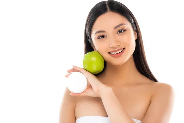 Morena mujer asiática sosteniendo manzana verde y crema cosmética cerca de la cara aislada en blanco - foto de stock