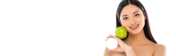 Panoramic concept of young asian woman holding cosmetic cream and green apple near face isolated on white — Stock Photo