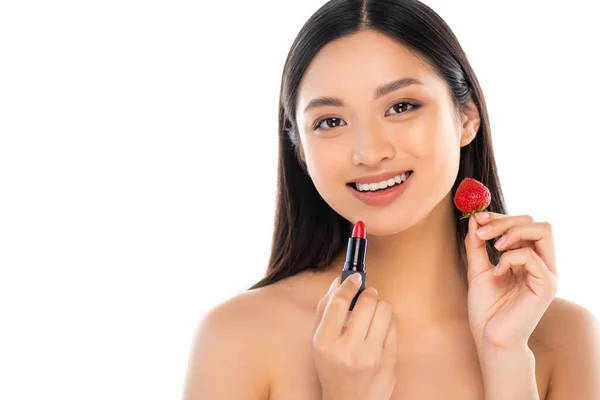 Mujer asiática desnuda mirando a la cámara mientras sostiene dulce fresa y lápiz labial rojo cerca de la cara aislada en blanco - foto de stock