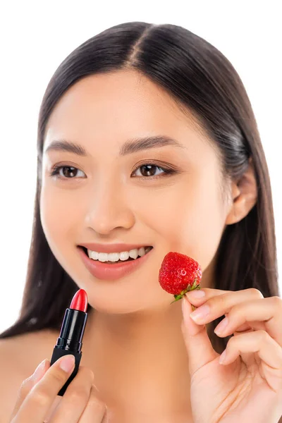 Morena mujer asiática mirando hacia otro lado mientras sostiene lápiz labial rojo y fresa madura cerca de la cara aislada en blanco - foto de stock