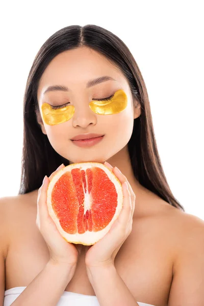 Brunette asian woman with golden eye patches on face holding half of ripe grapefruit with closed eyes isolated on white — Stock Photo