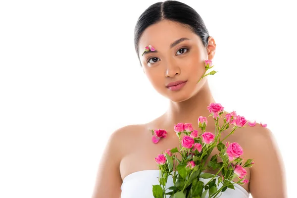 Sensual asian woman with flowers on face and petals on shoulder near pink roses isolated on white — Stock Photo