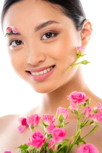 Young asian woman with floral decor on face looking at camera near pink roses isolated on white — Stock Photo