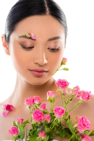 Mujer asiática desnuda con decoración floral en la cara cerca de rosas rosadas aisladas en blanco - foto de stock