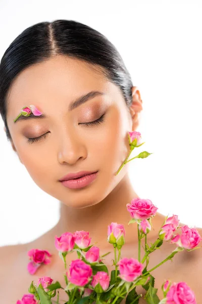 Junge asiatische Frau mit Knospen und Blütenblatt im Gesicht schließt die Augen in der Nähe rosa Rosen isoliert auf weiß — Stockfoto