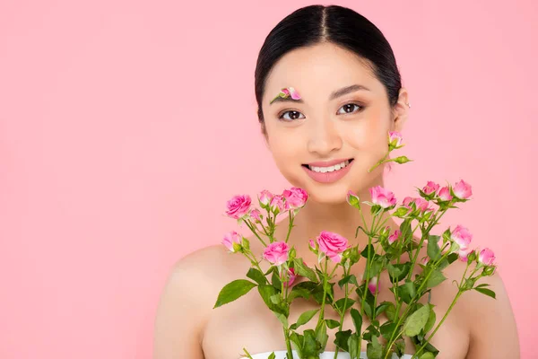 Brünette asiatische Frau mit Blumenschmuck im Gesicht hält winzige Rosen isoliert auf rosa — Stockfoto