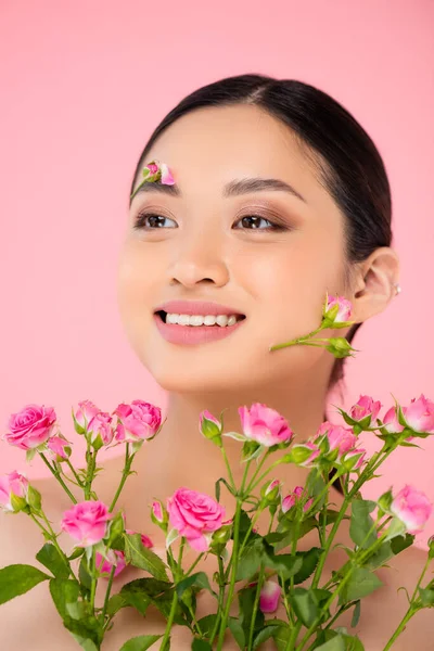 Jeune femme asiatique avec des fleurs sur le visage regardant loin près de minuscules roses isolées sur rose — Photo de stock