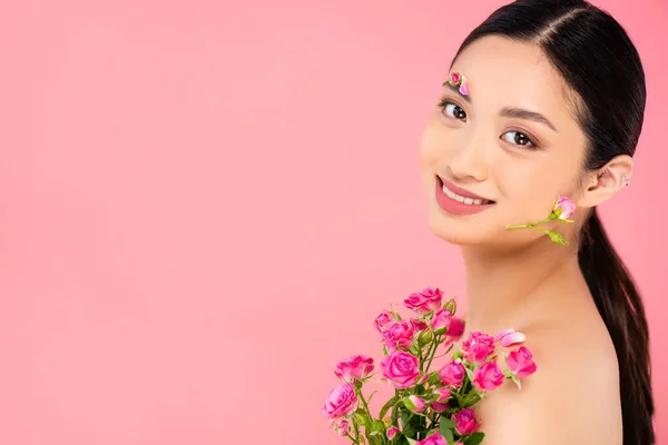 Femme asiatique nue avec décoration florale sur le visage et les pétales sur l'épaule tenant de minuscules roses isolées sur rose — Photo de stock