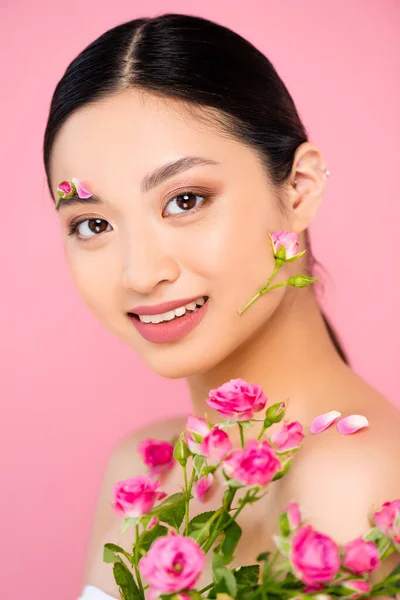 Brunette asian woman with buds and petals on face and shoulder, holding tiny roses isolated on pink — Stock Photo