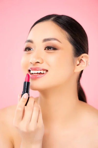 Nu asiatique femme regarder loin tout en appliquant rouge à lèvres isolé sur rose — Photo de stock