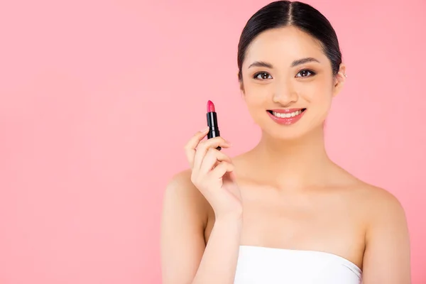 Morena mujer asiática sosteniendo lápiz labial y mirando a la cámara aislada en rosa - foto de stock