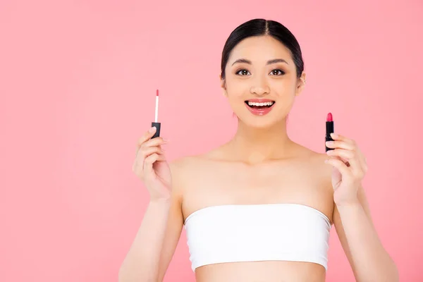 Excited asian woman holding lip gloss and lipstick while looking at camera isolated on pink — Stock Photo