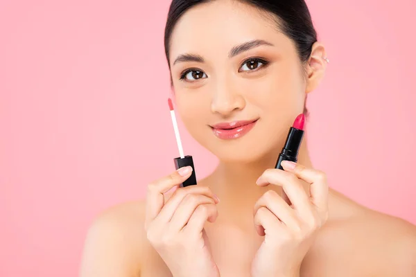 Mujer asiática desnuda sosteniendo lápiz labial y brillo de labios mientras mira a la cámara aislada en rosa - foto de stock