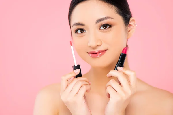Naked asian woman holding lipstick and lip gloss while looking at camera isolated on pink — Stock Photo